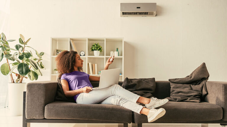 Mulher sentada no sofá de casa com o notebook no colo ligando o ar-condicionado através do controle remoto
