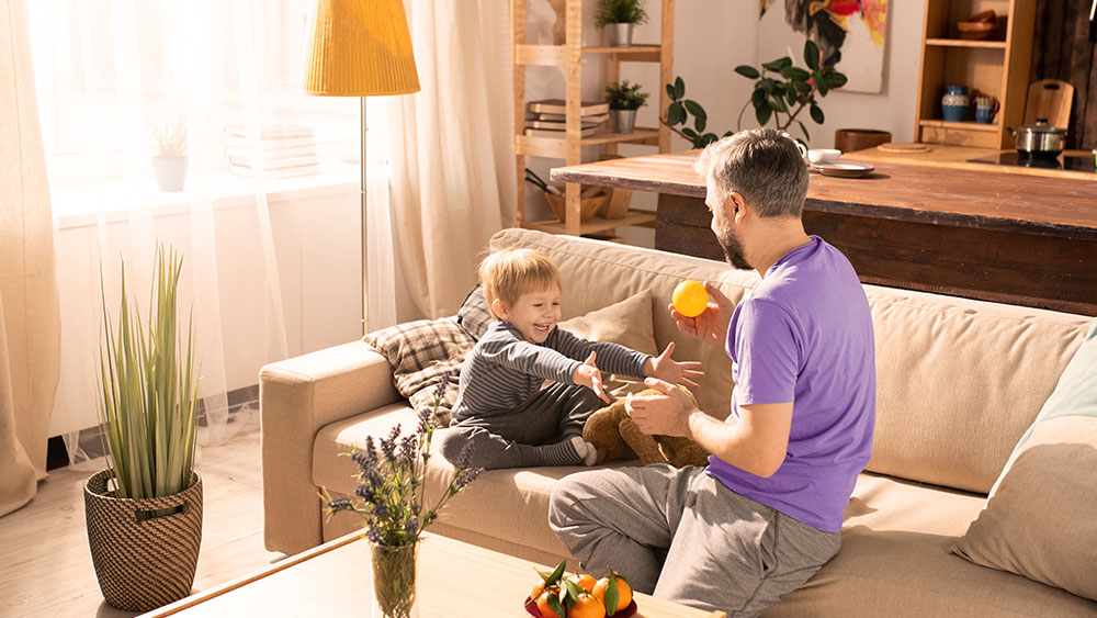 Pai e filho brincam com segurança em uma sala bem iluminada por luz natural, representando um lar com segurança elétrica e uso sustentável da energia.